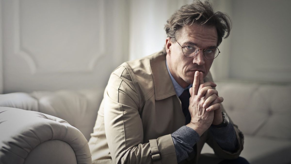 A man in thin round metal frames sitting on a pale sofa, hands folded