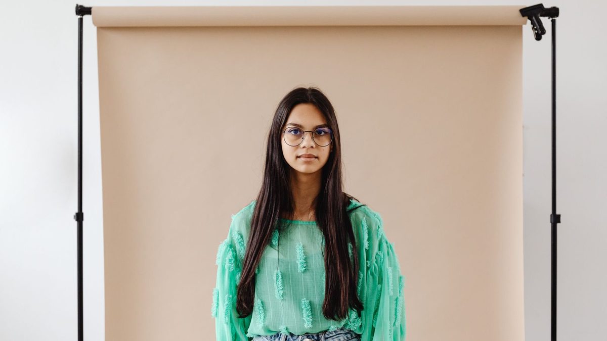 A woman in thin round frames standing in front of a pale studio backdrop